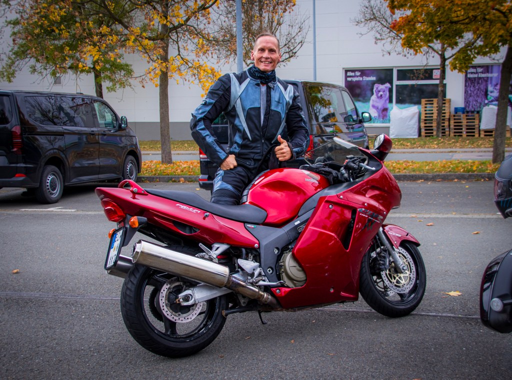 Ein Motorradfahrer steht neben seiner roten Sportmaschine auf einem Parkplatz, lächelt und gibt einen Daumen hoch. Herbstliche Bäume im Hintergrund.