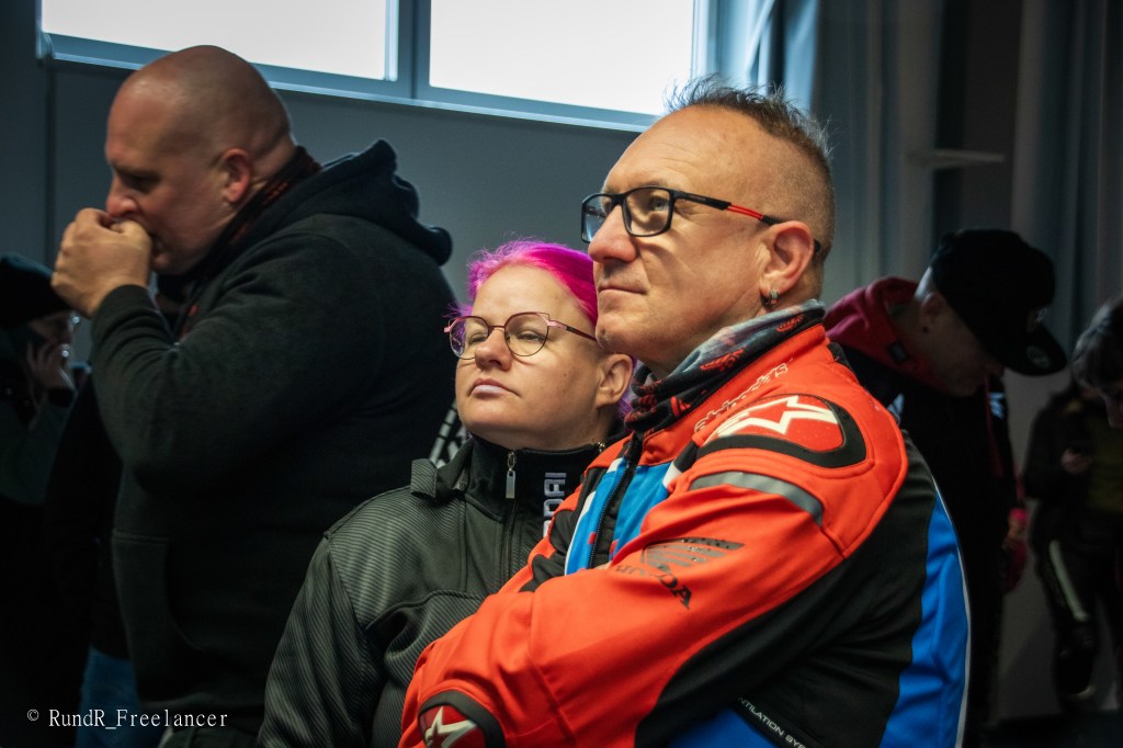 Zwei Personen, eine mit rosa Haaren und Brille, die andere mit dreieckiger Frisur und in einer motorisierten Jacke, hören aufmerksam zu. Im Hintergrund sind weitere Personen sichtbar.