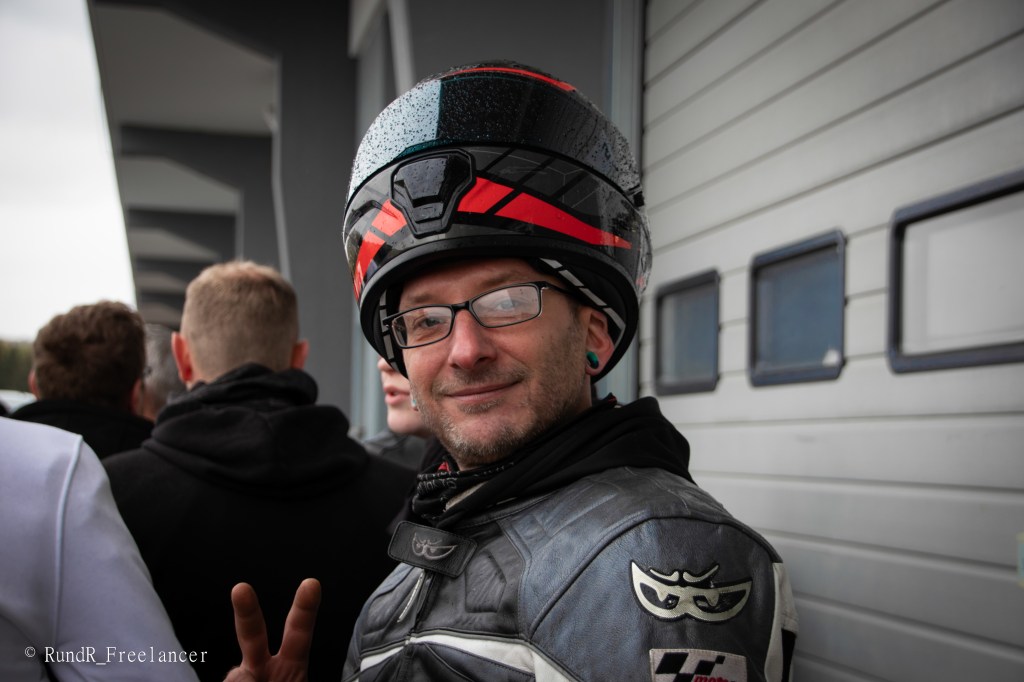 Ein Motorradfahrer mit einem Helm und einer Jacke schaut lächelnd in die Kamera und macht ein Peace-Zeichen, während er von anderen Personen umgeben ist.