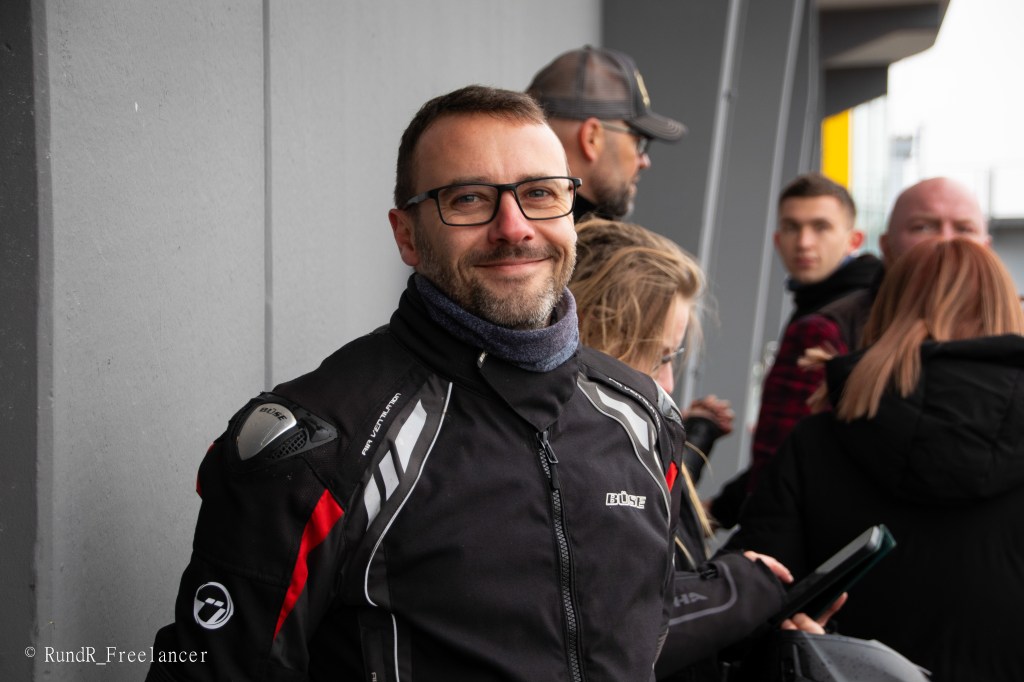 Ein lächelnder Mann in Motorradkleidung steht vor einer grauen Wand, umgeben von anderen Personen.