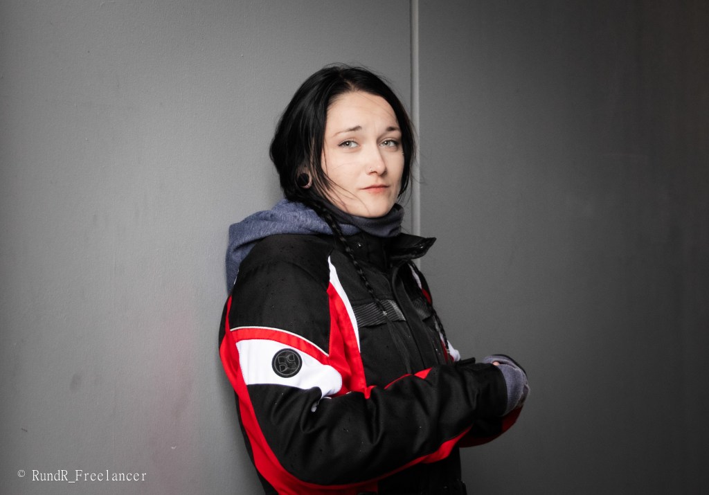 Eine Person mit langen, dunklen Haaren, gekleidet in eine schwarze Jacke mit roten und weißen Akzenten, steht an einer grauen Wand und schaut direkt in die Kamera.