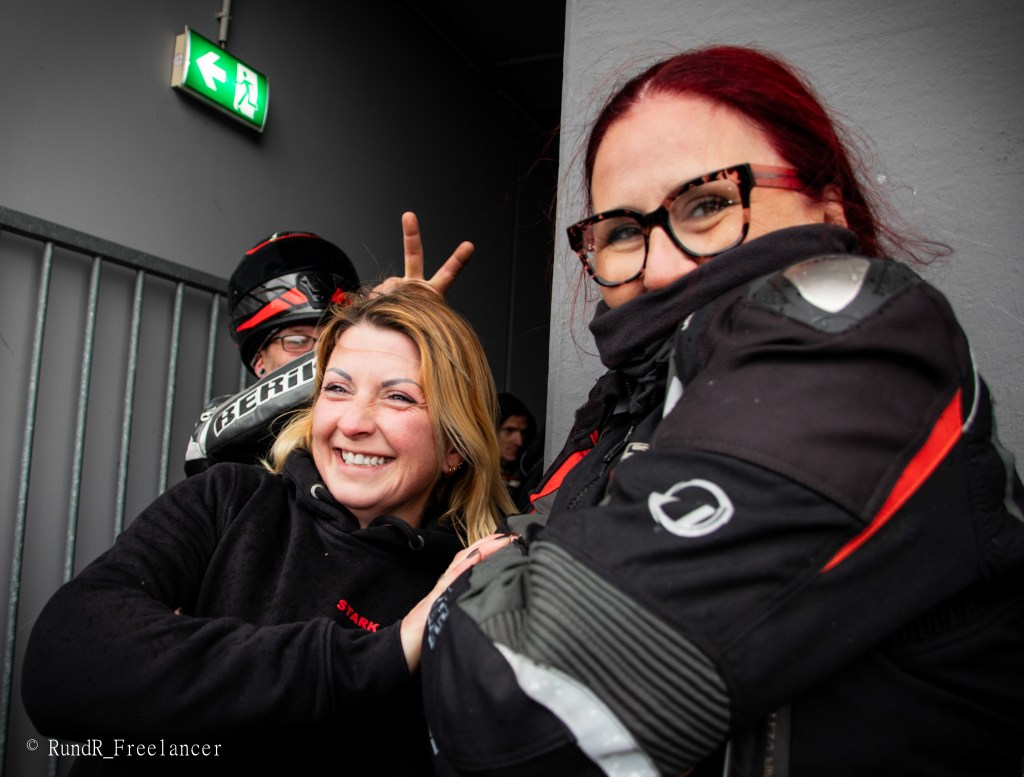 Zwei lächelnde Frauen in Motorradkleidung, die eng beieinander stehen. Eine Frau hat blonden Haar und die andere hat rot gefärbtes Haar. Im Hintergrund ist eine Person mit einem Motorradhelm, die mit den Fingern ein freches Zeichen macht.