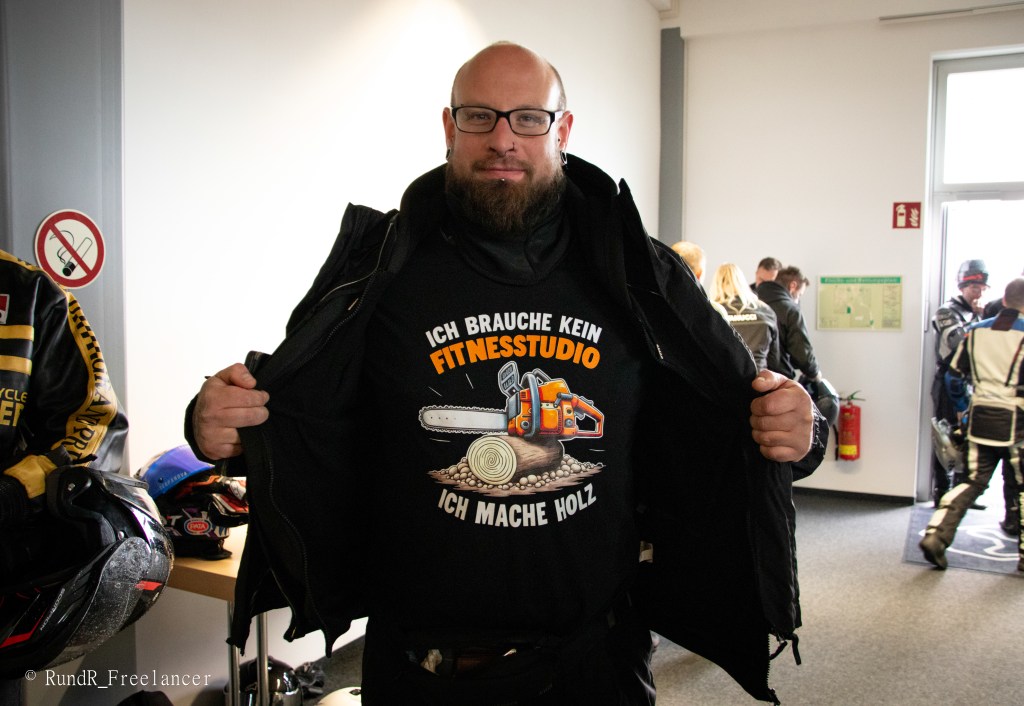 Ein Mann steht in einem Raum und präsentiert ein T-Shirt mit der Aufschrift 'Ich brauche kein Fitnessstudio, ich mache Holz'.