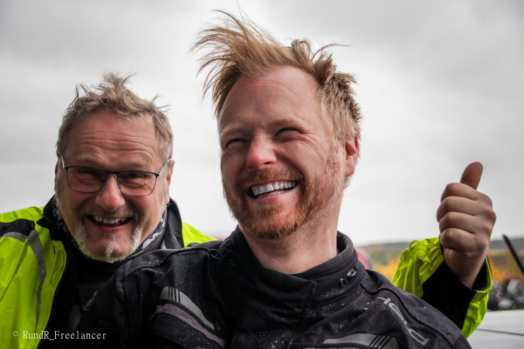 Zwei Männer lächeln und posieren fröhlich in Motorradbekleidung, während der Wind ihre Haare zerzaust. Einer zeigt Daumen hoch.