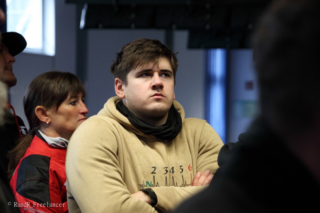 Ein junger Mann mit verschränkten Armen schaut aufmerksam nach vorne, während eine Frau in der Nähe im Hintergrund steht. Der Mann trägt einen beigen Pullover und einen schwarzen Schal.