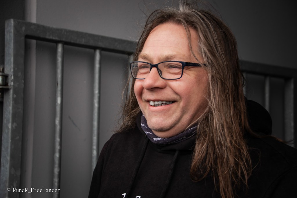 Ein lächelnder Mann mit langen, lockigen Haaren und Brille steht vor einem Zaun.