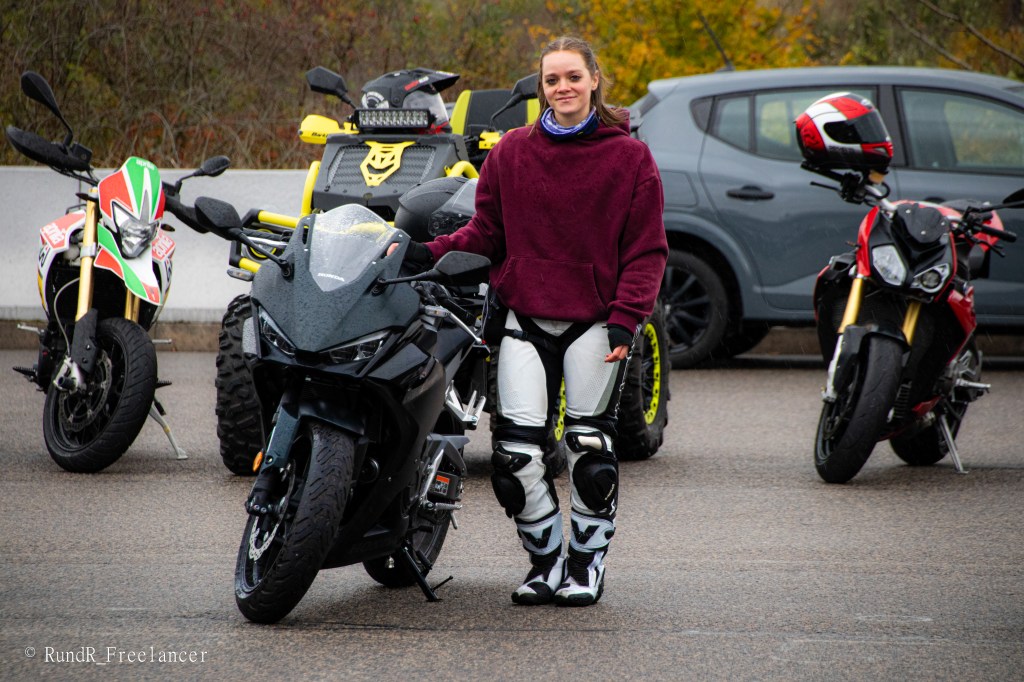 Eine Motorradfahrerin steht neben ihrem Sportmotorrad, umgeben von verschiedenen anderen Motorrädern und einem Auto im Hintergrund.