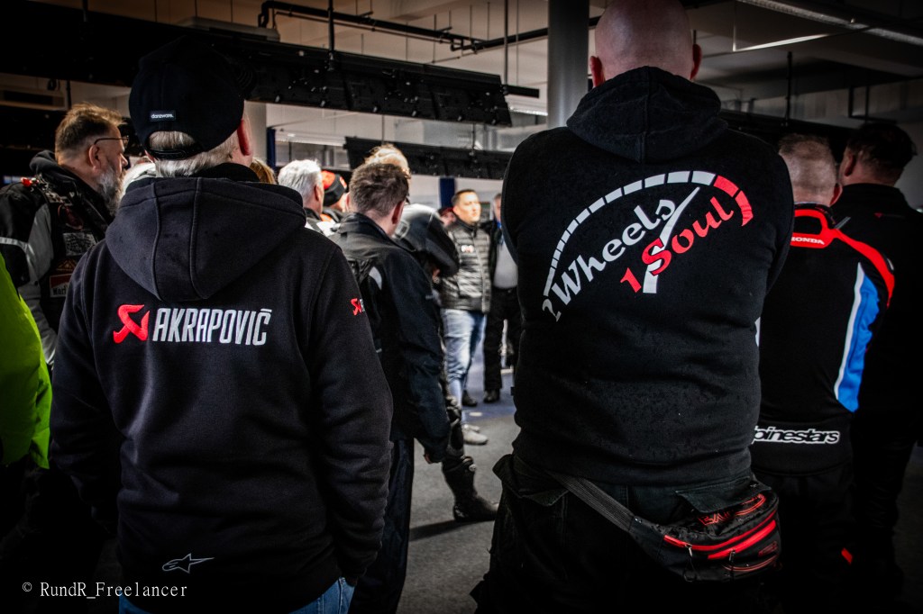 Gruppe von Motorradfahrern, die sich um einen Redner versammelt hat, mit verschiedenen Jacken und Logos, darunter 'Akrapovič' und 'Wheels 1 Soul'.