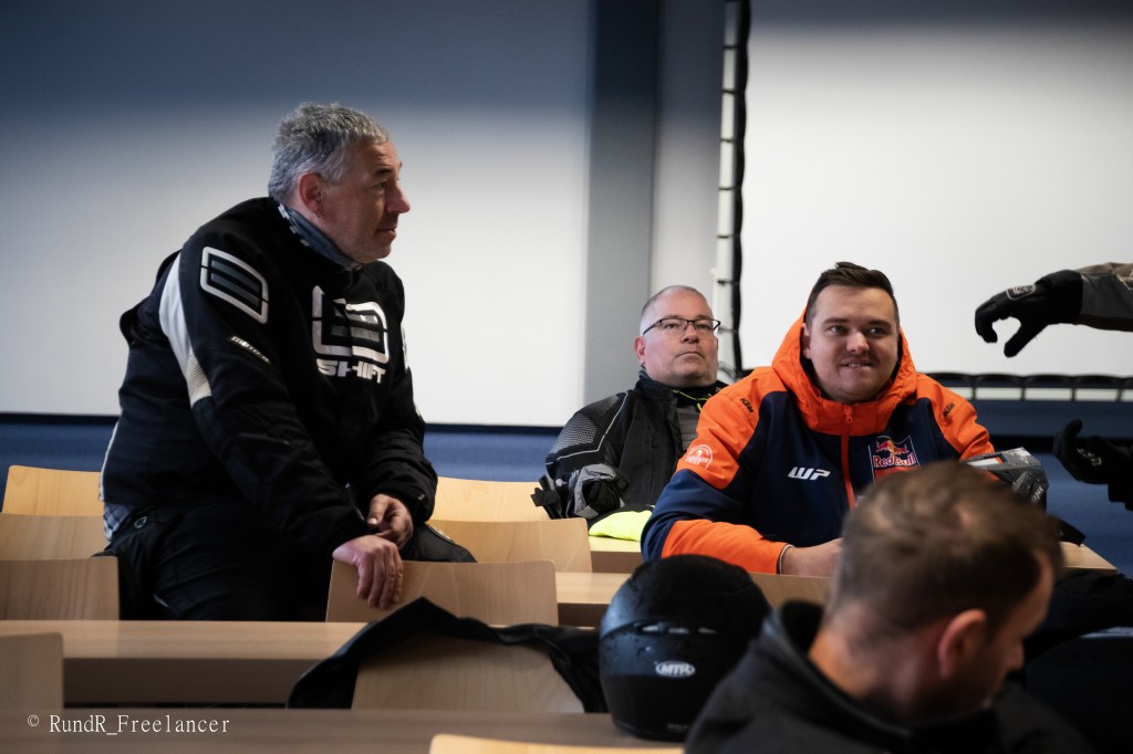 Drei Männer in Motorradkleidung sitzen in einem Seminarraum mit Holztischen und hellen Wänden.