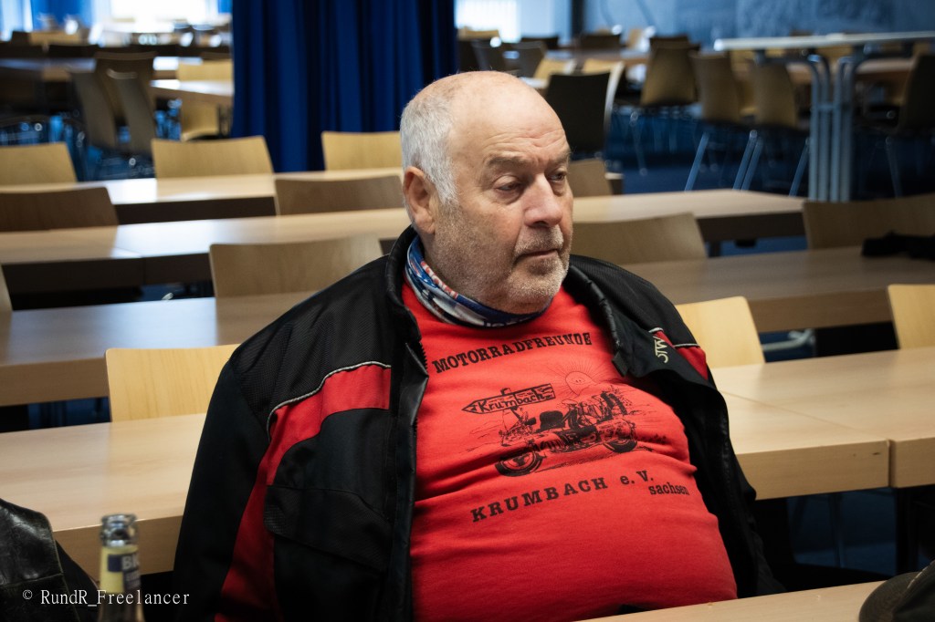 Ein älterer Mann mit Glatze sitzt in einem Raum voller Tische und Stühle, trägt ein rotes T-Shirt mit Motorradmotiv und eine schwarze Jacke.