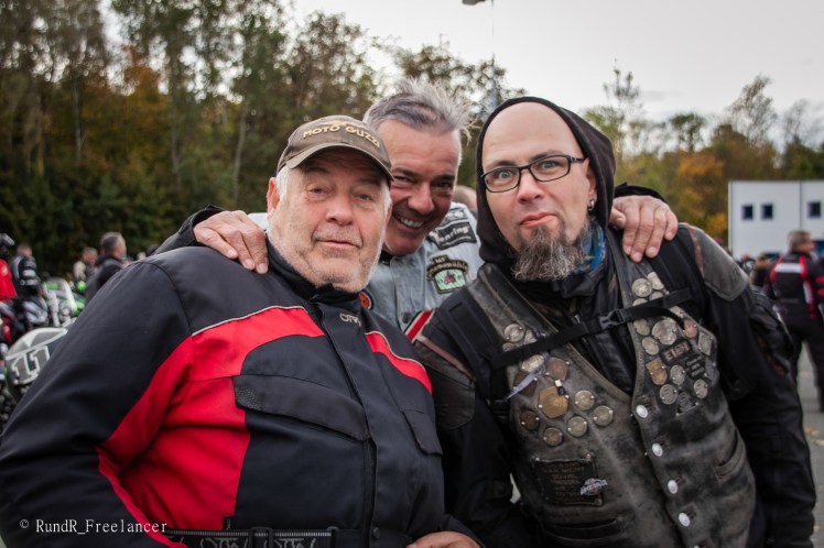 Drei Männer posieren freundlich für ein Gruppenfoto, während sie Motorradjacken tragen. Im Hintergrund sind weitere Motorradfahrer und Motorräder zu sehen.
