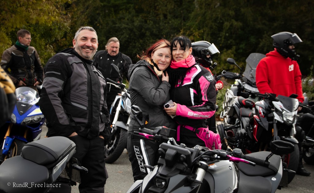 Eine Gruppe von Motorradfahrern versammelt sich mit mehreren Motorrädern im Hintergrund. Zwei Frauen umarmen sich lächelnd, während ein Mann in der Nähe grinst.