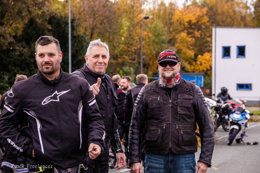 Eine Gruppe von Motorradfahrern posiert lächelnd in einer Parkumgebung, umgeben von Motorrädern und herbstlichen Bäumen.