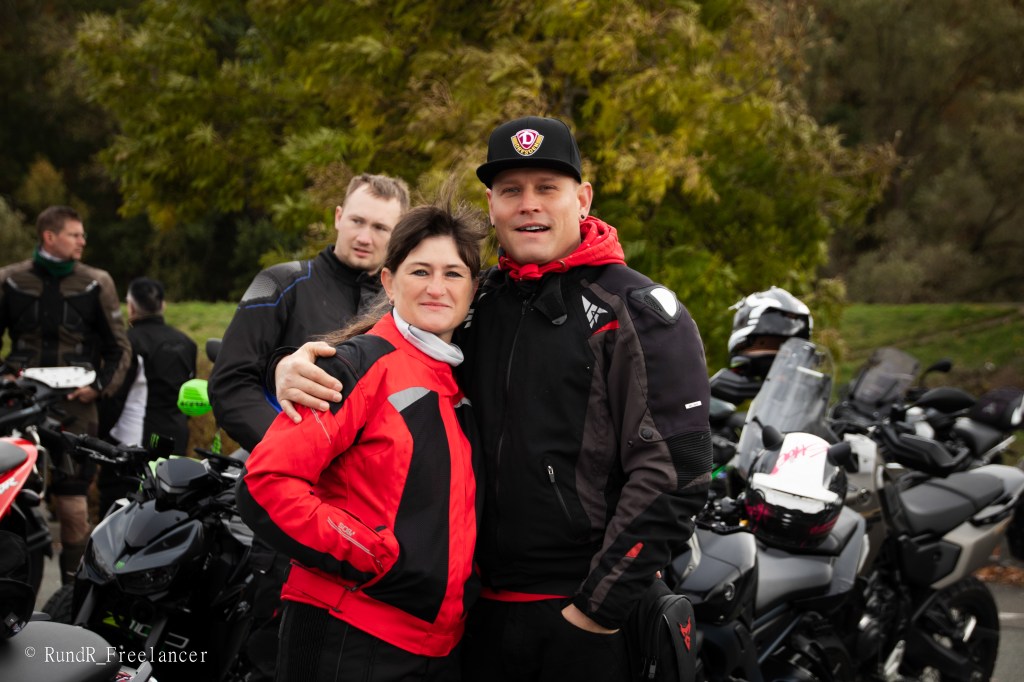Ein Paar posiert lächelnd vor einer Gruppe von Motorrädern, während weitere Motorradfahrer im Hintergrund stehen.