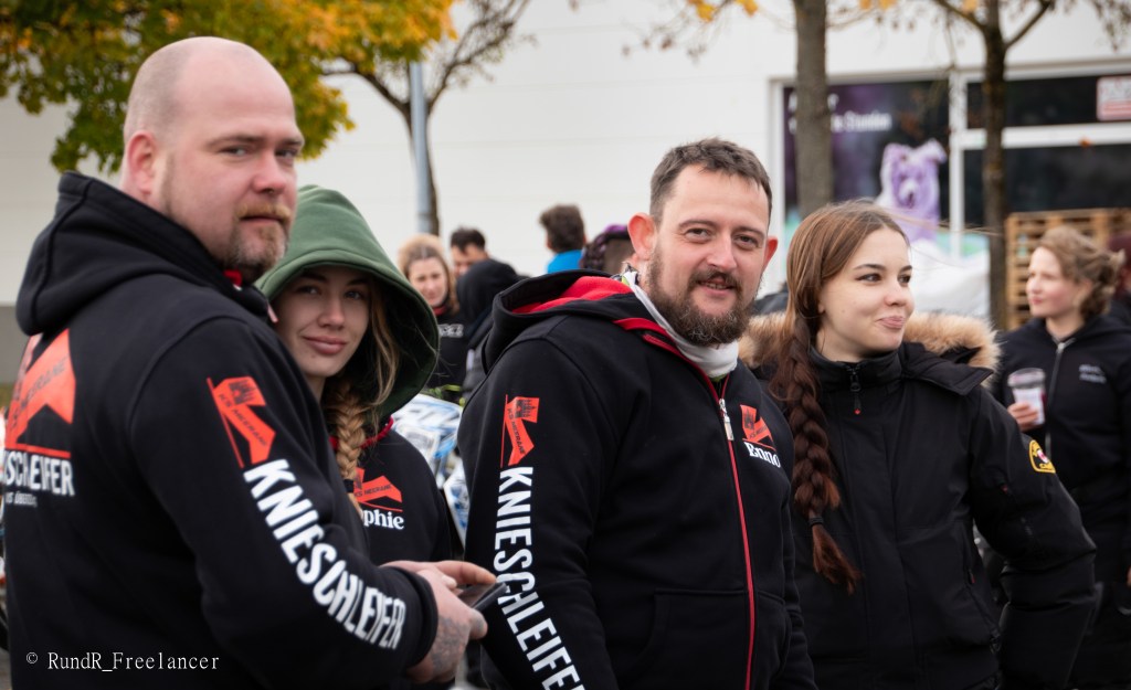 Eine Gruppe von vier Personen in Freizeitkleidung, lächelnd und sich unterhaltend, stehen draußen auf einem Event, mit einigen Bäumen im Hintergrund.