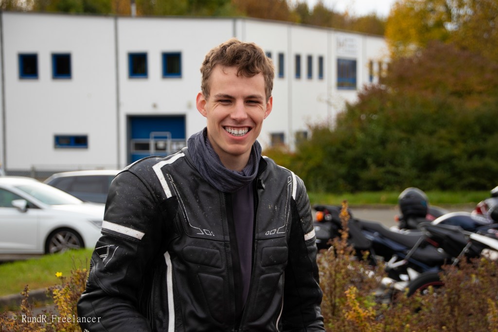 Ein junger Mann in einer Motorradjacke lächelt auf einem Parkplatz mit mehreren Motorrädern im Hintergrund.