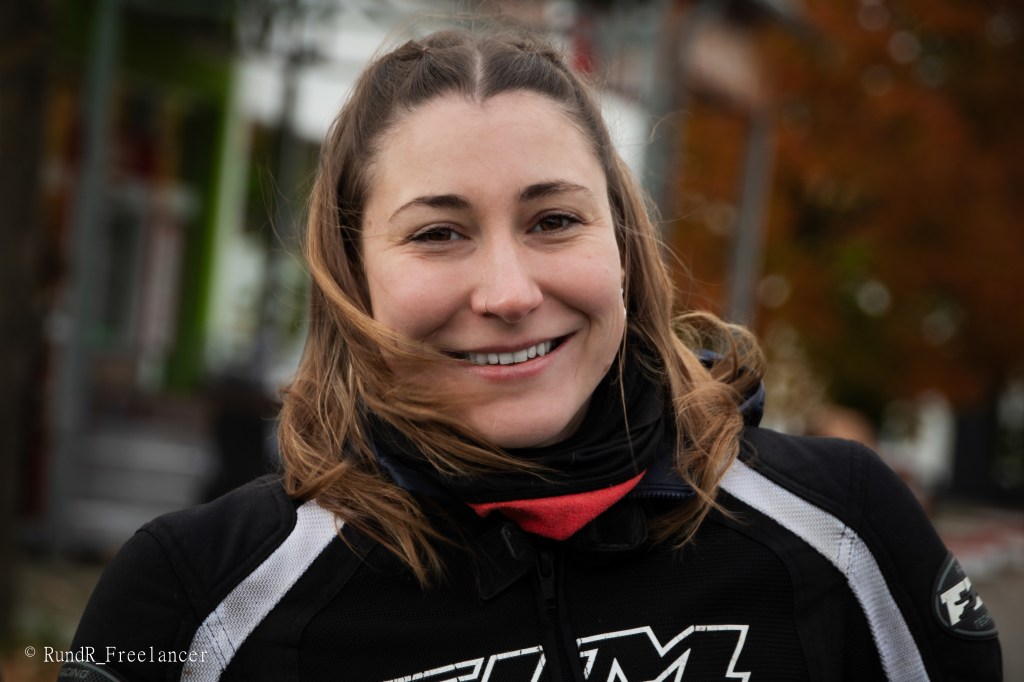 Eine lächelnde Frau mit langen, lockigen Haaren trägt eine Motorradjacke. Der Hintergrund zeigt unscharfe Farben von Bäumen und Gebäuden.