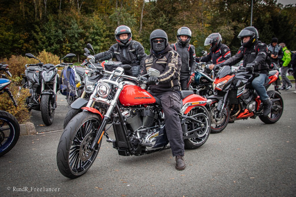 Alter Mann auf einer roten Harley-Davidson-Motorrad, umgeben von anderen Motorradfahrern in Schutzkleidung, die an einem Parkplatz stehen. Hintergrund zeigt Bäume und Umgebung.
