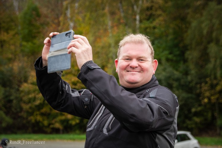 Ein Mann in schwarzer Motorradbekleidung lächelt und hält ein Smartphone im Hochformat in der Hand, während er ein Foto macht.