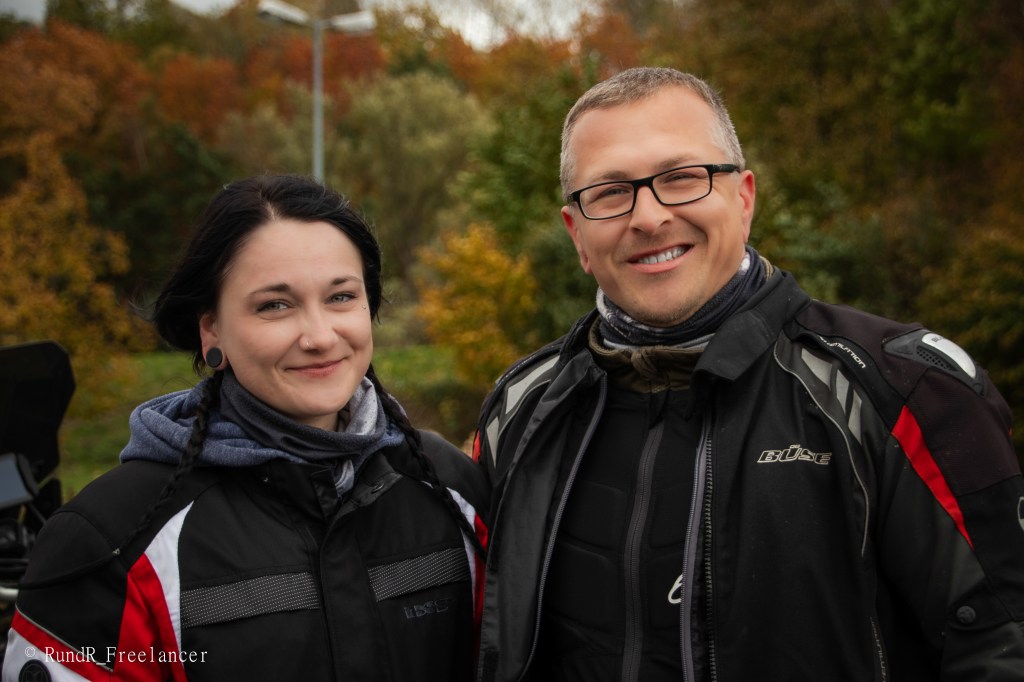 Zwei Motorradfahrer lächeln vor einer herbstlichen Hintergrundlandschaft mit buntem Laub.