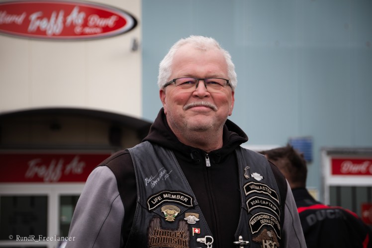 Ein lächelnder Mann mit Brille und grauen Haaren trägt eine schwarze Weste mit verschiedenen Abzeichen, steht vor einem Gebäude mit einem Schild im Hintergrund.