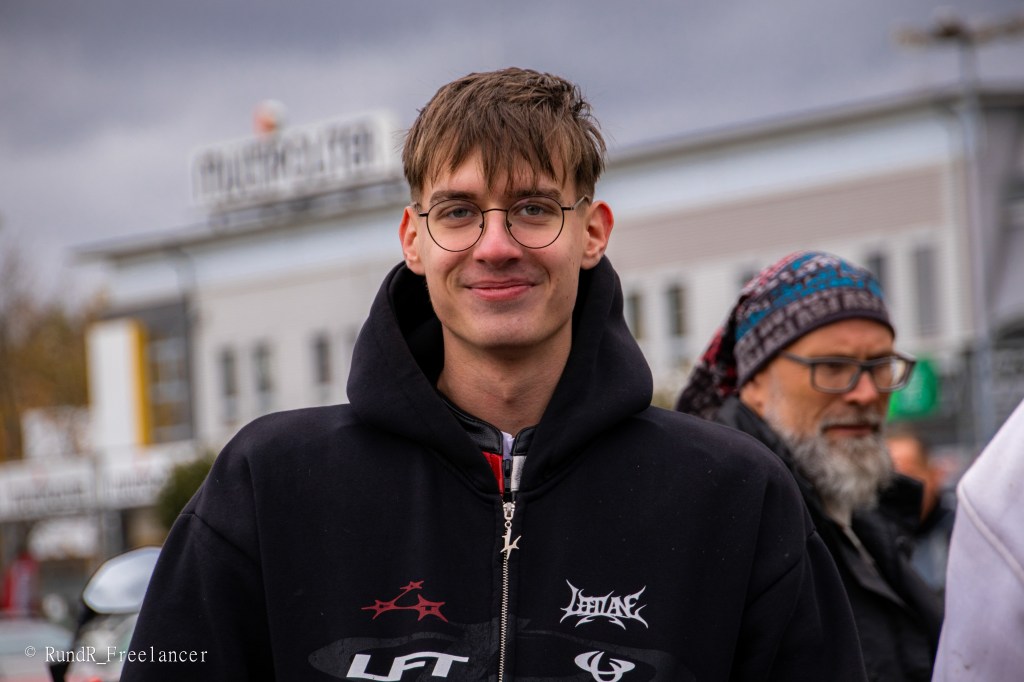 Ein junger Mann mit Brille und einem lächelnden Gesicht, der einen schwarzen Kapuzenpullover trägt. Im Hintergrund sind unscharfe Personen und ein Gebäude sichtbar.