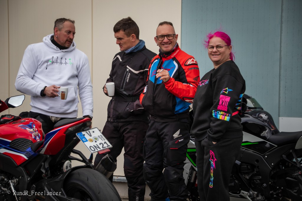 Vier Motorradfahrer stehen vor ihren Motorrädern und unterhalten sich. Sie halten Tassen in den Händen und tragen Motorradbekleidung.