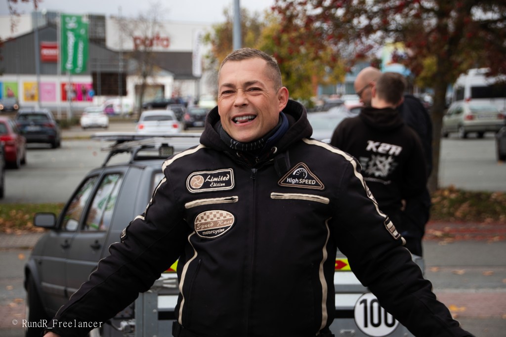 Ein lachender Mann in einem Motorradjacke steht vor einem Parkplatz, umgeben von Autos und Bäumen im Herbst. Im Hintergrund sind einige Personen und ein Einkaufszentrum zu sehen.
