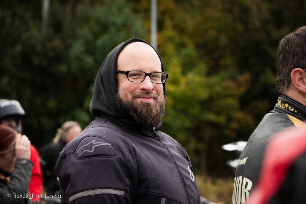 Ein lächelnder Mann mit Brille und einem schwarzen Kapuzenpullover, umgeben von anderen Personen in einem natürlichen, grünen Umfeld.