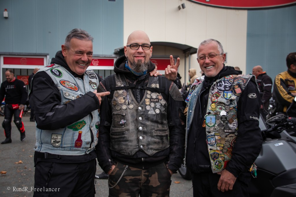 Drei Motorradfahrer posieren lächelnd vor einem Gebäude, während sie ihre Jacken mit verschiedenen Abzeichen und Patches tragen.