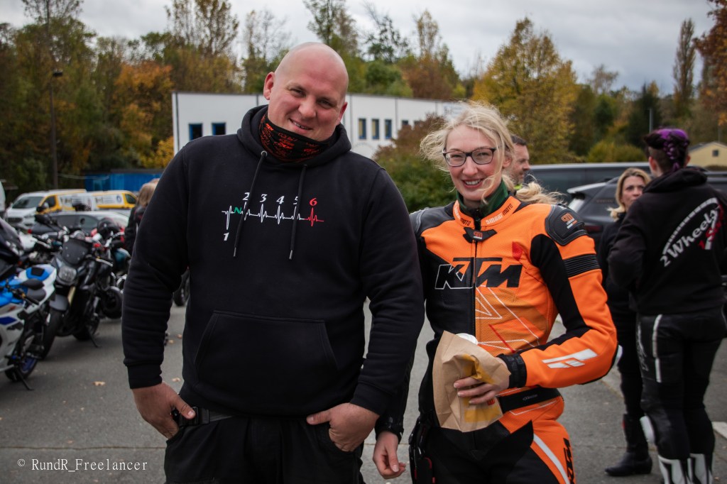 Ein Mann in einem schwarzen Hoodie und eine Frau in einer orangefarbenen Motorradjacke stehen lächelnd nebeneinander auf einem Parkplatz, umgeben von Motorrädern und herbstlicher Landschaft.