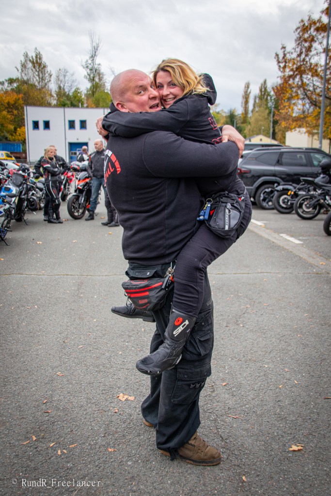 Ein muskulöser Mann hebt eine lächelnde Frau in einem Motorrad-Outfit an, während sie auf einem Parkplatz stehen. Im Hintergrund sind mehrere Motorräder und andere Personen zu sehen.