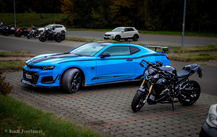 Ein blauer Chevrolet Camaro und ein schwarzes Motorrad stehen auf einem Parkplatz, umgeben von anderen Fahrzeugen.