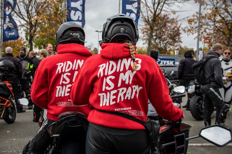 Zwei Motorradfahrerinnen in roten Hoodies mit dem Aufdruck 'RIDING IS MY THERAPY' stehen nebeneinander, umgeben von einer Gruppe von Menschen auf einem Motorrad-Event.
