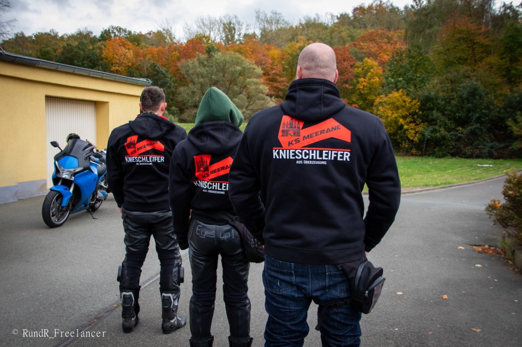 Drei Personen mit Motorradjacken stehen mit dem Rücken zur Kamera, während sie auf eine landschaftlich reizvolle Umgebung blicken. Im Hintergrund steht ein Motorrad und Bäume zeigen herbstliche Farben.