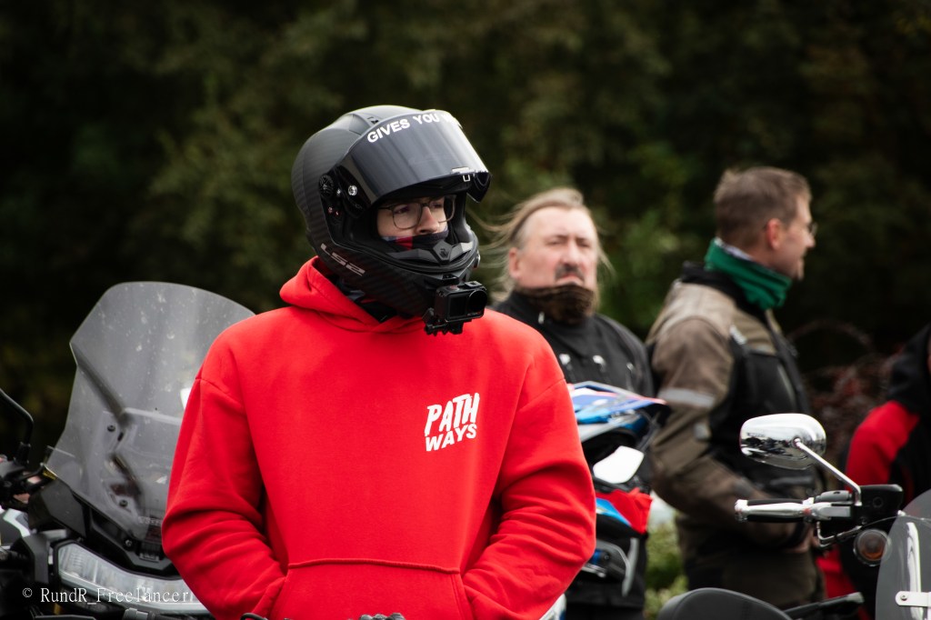 Ein Motorradfahrer in einem roten Hoodie steht in der Menge, trägt einen schwarzen Helm mit der Aufschrift 'GIVES YOU' und schaut nachdenklich. Im Hintergrund sind weitere Motorradfahrer und Motorräder zu sehen.