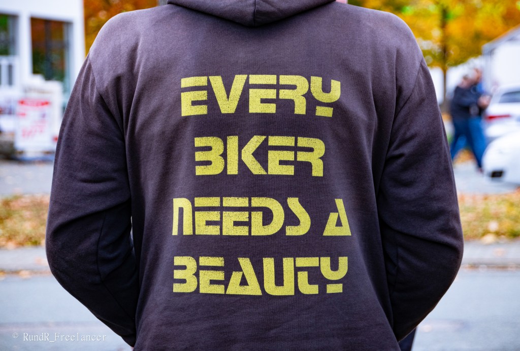 Ein Mann trägt einen dunkelgrauen Hoodie mit dem Aufdruck "EVERY BIKER NEEDS A BEAUTY" auf dem Rücken. Im Hintergrund sind herbstliche Bäume und Autos zu sehen.