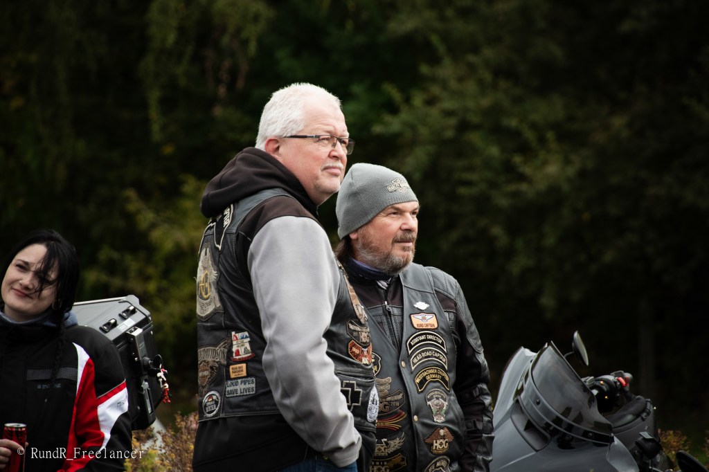 Zwei Männer in Motorradjacken stehen in der Natur und sehen in die Ferne, während eine Frau im Hintergrund lächelt.