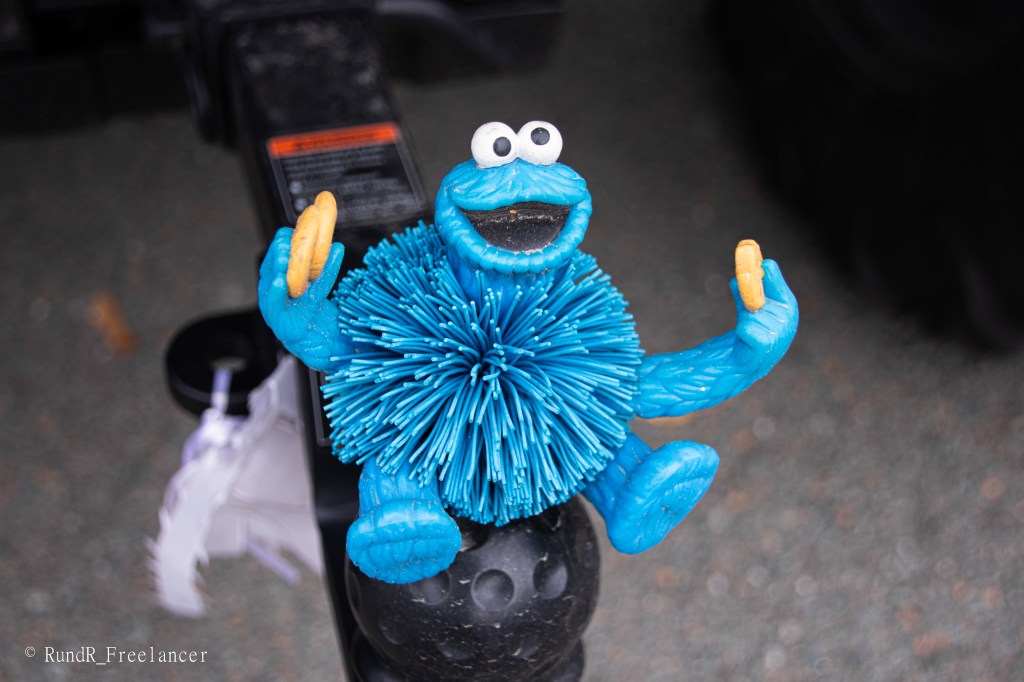 Eine blaue Kunststofffigur mit großen Augen, die wie ein Monster aussieht und einen Keks in jeder Hand hält. Die Figur hat einen runden, bauschigen Körper und sitzt auf einer Anhängerkupplung.