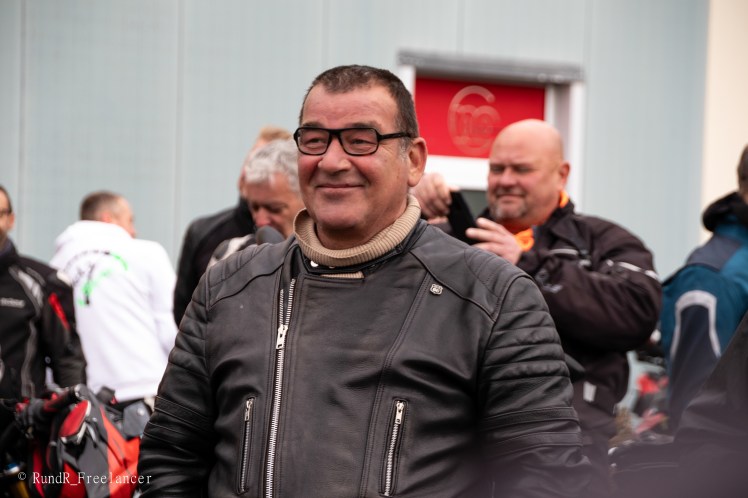 Ein lächelnder Mann in einer schwarzen Motorradlederjacke mit Brille, umgeben von anderen Motorradfahrern vor einem Gebäude.