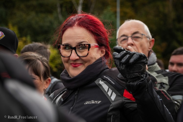 Eine Person mit roten Haaren und Brille, die in Motorradkleidung gekleidet ist, lächelt und zeigt mit der Hand. Im Hintergrund sind weitere Personen zu sehen.
