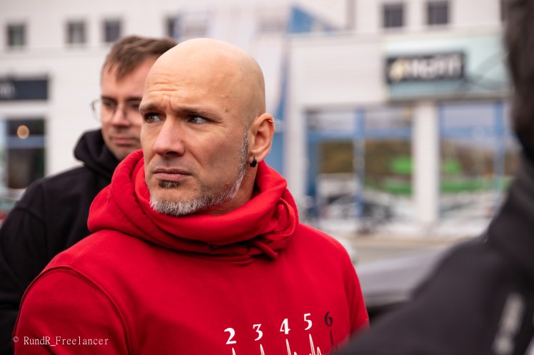 Mann mit Glatze und Bart in einem roten Hoodie, der nachdenklich aussieht. Im Hintergrund sind Menschen und Gebäude zu sehen.