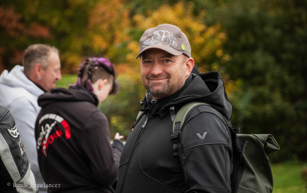 Ein Mann mit dunkler Jacke und einer grauen Kappe lächelt in die Kamera vor einer herbstlichen Kulisse.