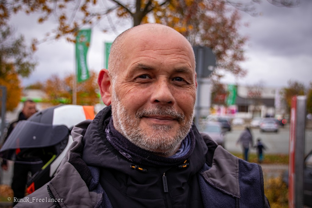 Ein älterer Mann mit kurzen, grauen Haaren und einem Bart lächelt in die Kamera. Er trägt eine schwarze Jacke und hat eine Motorradhelmkappe in der Nähe.