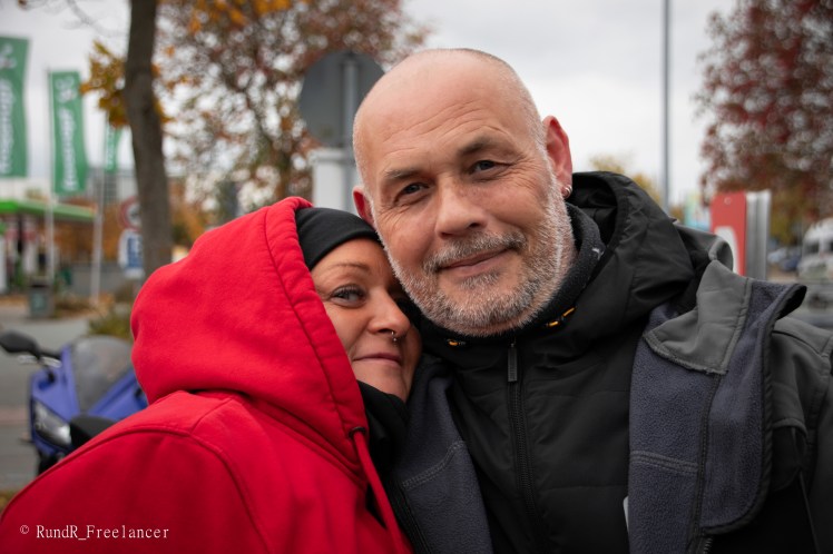 Ein Paar umarmt sich in einer urbanen Umgebung, beide lächeln in die Kamera. Die Frau trägt einen roten Kapuzenpullover, während der Mann einen schwarzen Mantel anhat.