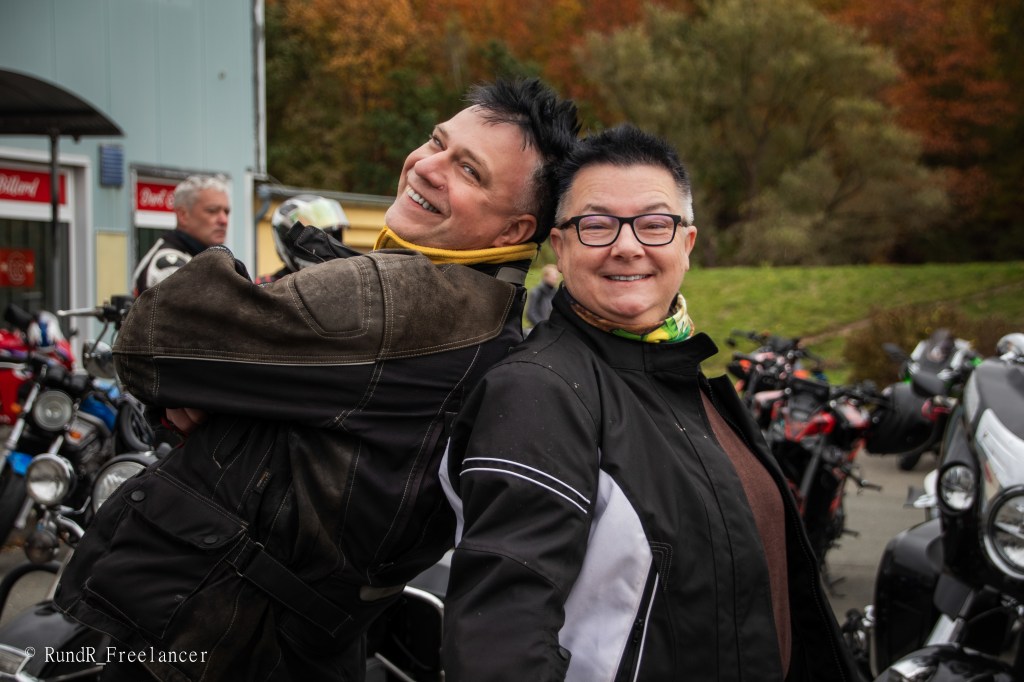 Zwei Motorradfahrer lächeln und stehen Rücken an Rücken vor einer Reihe von Motorrädern.