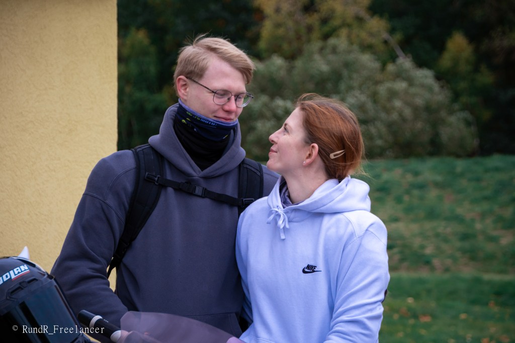 Ein junges Paar steht nahe beieinander und schaut sich an, während sie an einem Motorrad stehen. Der Mann trägt eine Brille und einen Schal, die Frau trägt einen grauen Hoodie.
