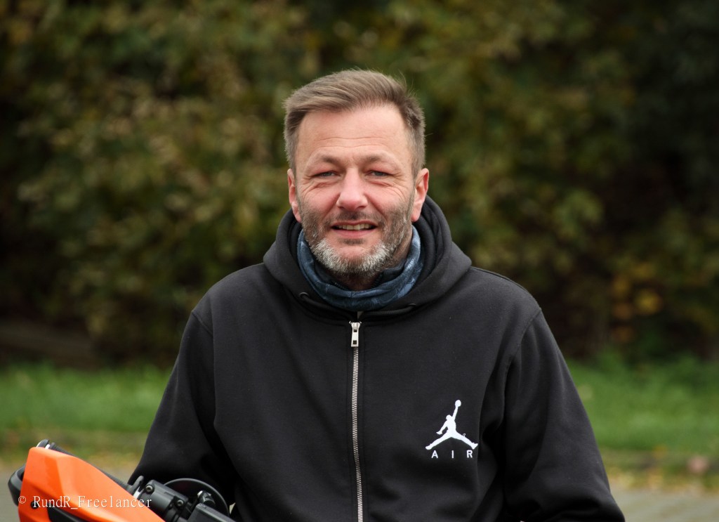 Ein lächelnder Mann mit Bart trägt einen schwarzen Kapuzenpullover und einen Schal, steht vor einem unscharfen, grünen Hintergrund.