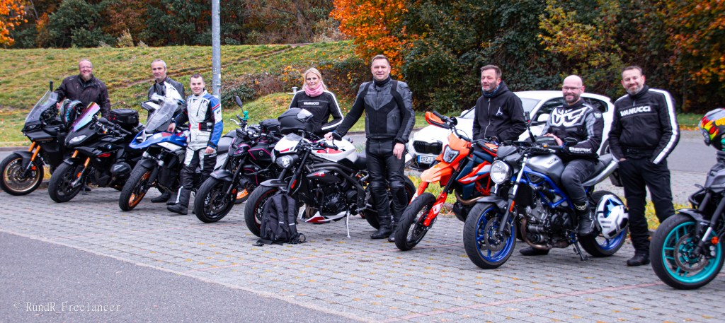Eine Gruppe von Motorradfahrern steht in voller Motorradmontur neben ihren Motorrädern auf einem Asphaltparkplatz, umgeben von herbstlichen Bäumen.