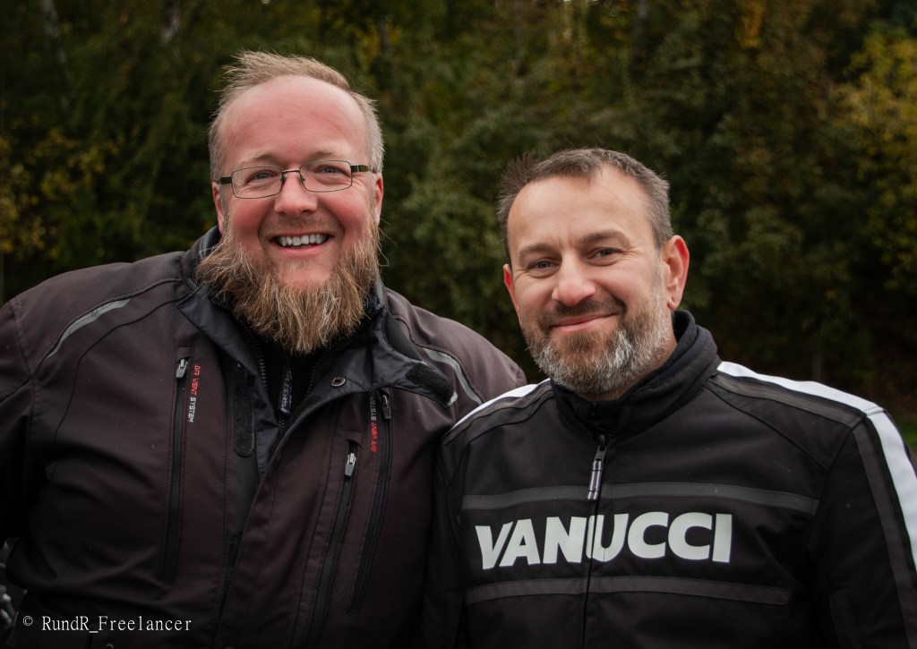 Zwei Männer mit Bart, die in Motorradjacken lächeln, stehen im Freien vor einer grünen Naturkulisse.